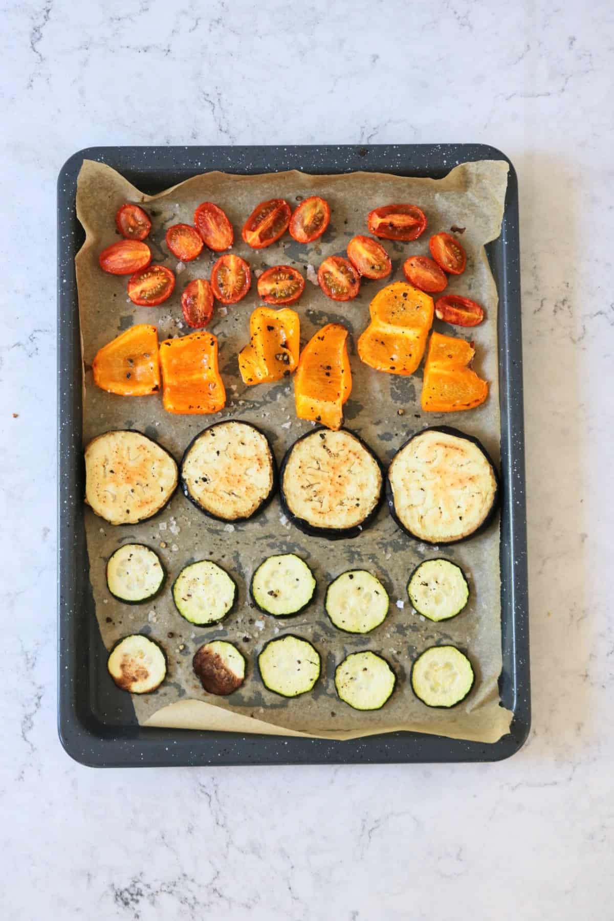 A baking tray with rows of roasted cherry tomatoes, orange pepper slices, aubergine slices and courgette slices