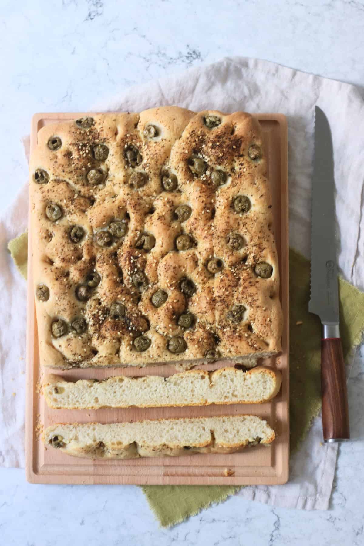 Sliced Green Olive and Za'atar Focaccia on a wooden chopping board with a large bread knife