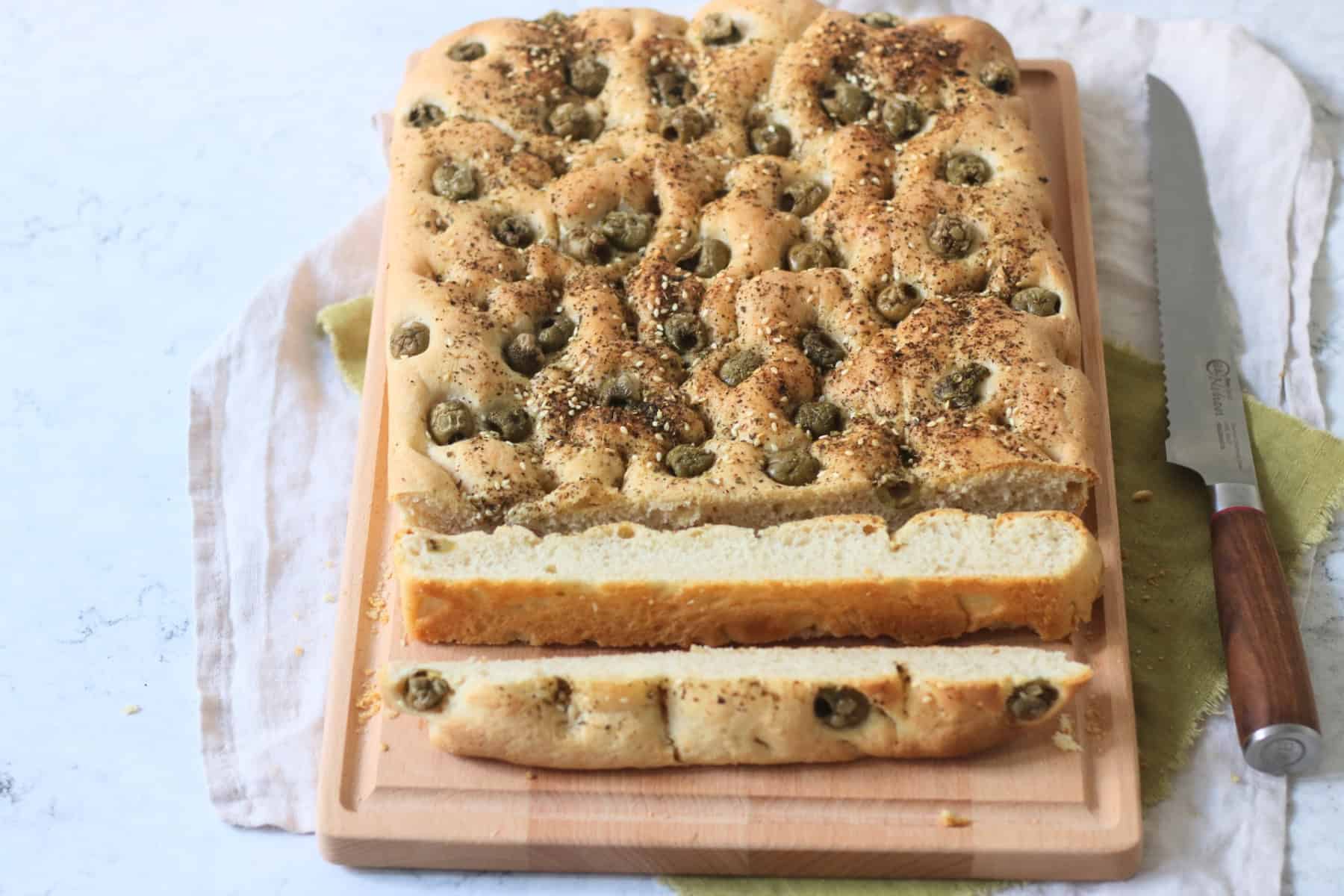 Sliced up Green Olive and Za'atar Focaccia on a wooden chopping board with a large bread knife