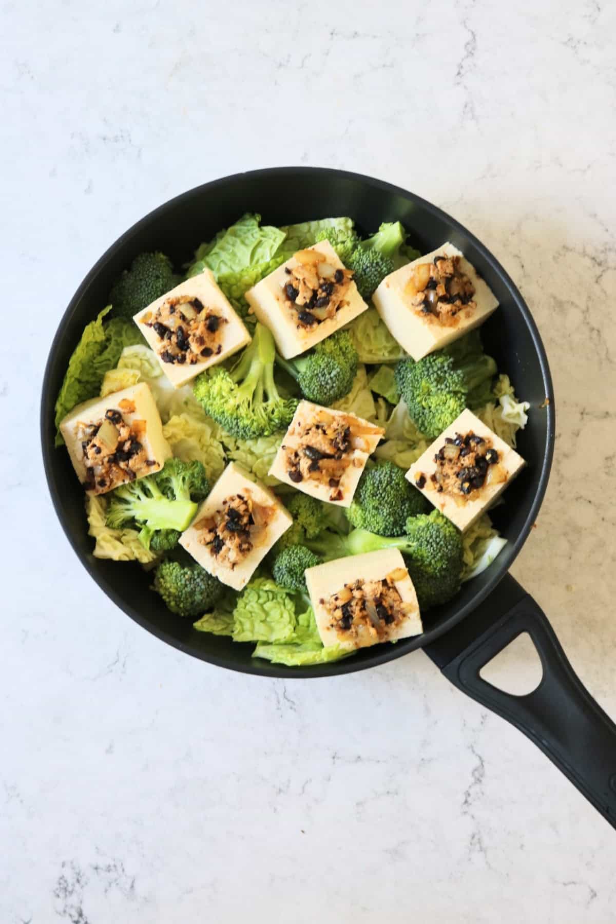 Black bean stuffed tofu nestled amongst cabbage and broccoli in a large frying pan ready to be braised