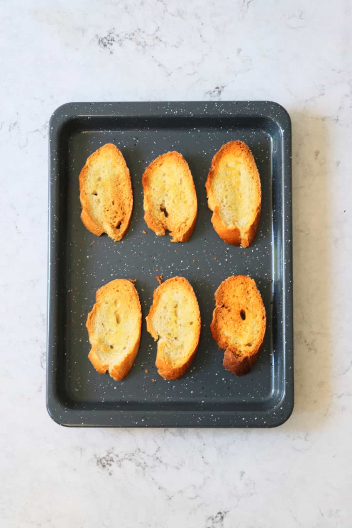 6 slices of toasted baguette on a grey baking sheet.