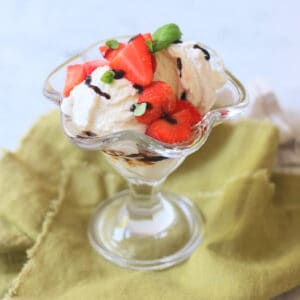 A Vegan Strawberry and Balsamic Ice Cream Sundae made up of layers of balsamic glaze, dairy free ice cream, macerated strawberries and basil leaves served in an ice cream sundae glass on top of a green napkin.