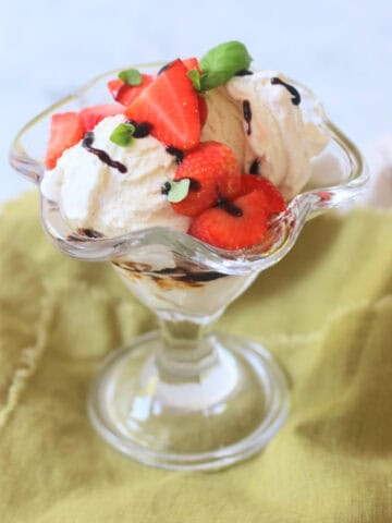 A Vegan Strawberry and Balsamic Ice Cream Sundae made up of layers of balsamic glaze, dairy free ice cream, macerated strawberries and basil leaves served in an ice cream sundae glass on top of a green napkin.