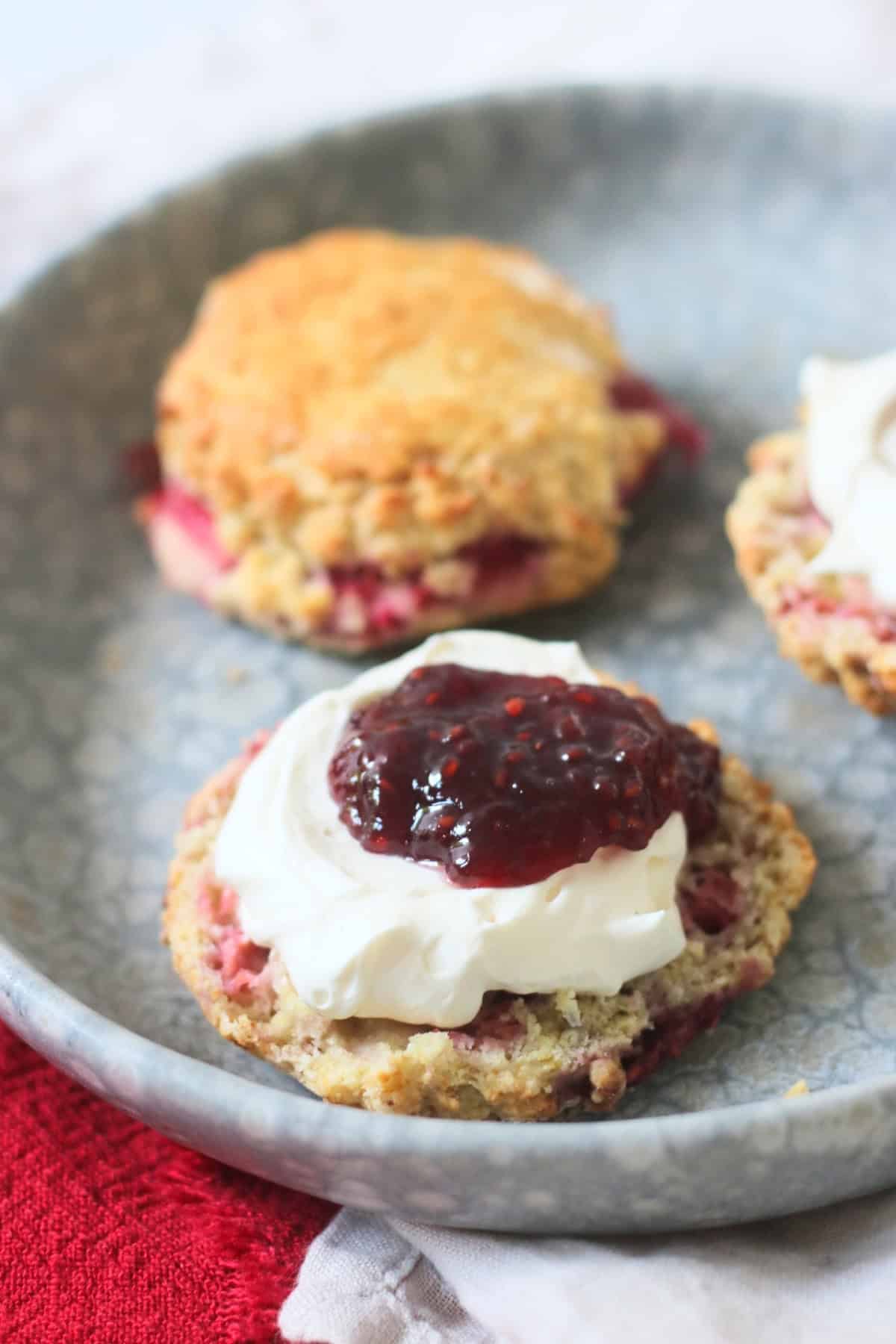 A Vegan Raspberry and Lemon Scone on a plate topped with vegan cream and raspberry jam