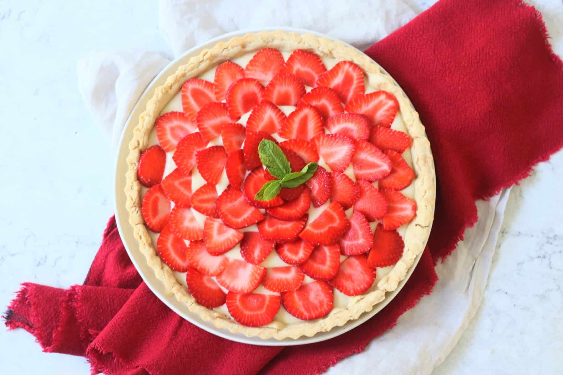 A whole Vegan Strawberry Tart (Vegan Tarte Aux Fraises) made up of a crisp pastry case, vegan creme patisserie and slices of strawberry topped with a couple of mint leaves.