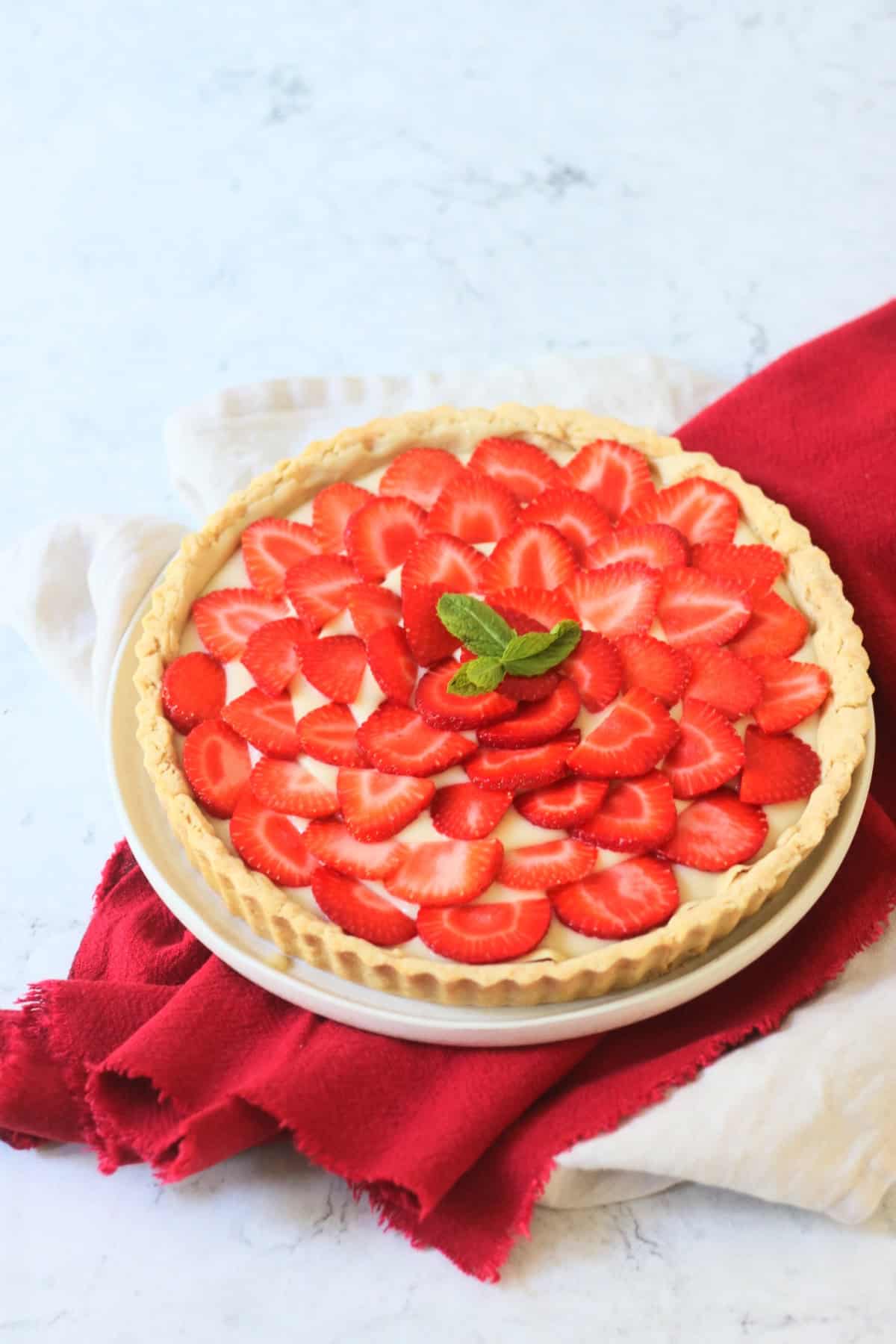 A whole Vegan Strawberry Tart (Vegan Tarte Aux Fraises) made up of a crisp pastry case, vegan creme patisserie and slices of strawberry topped with a couple of mint leaves.