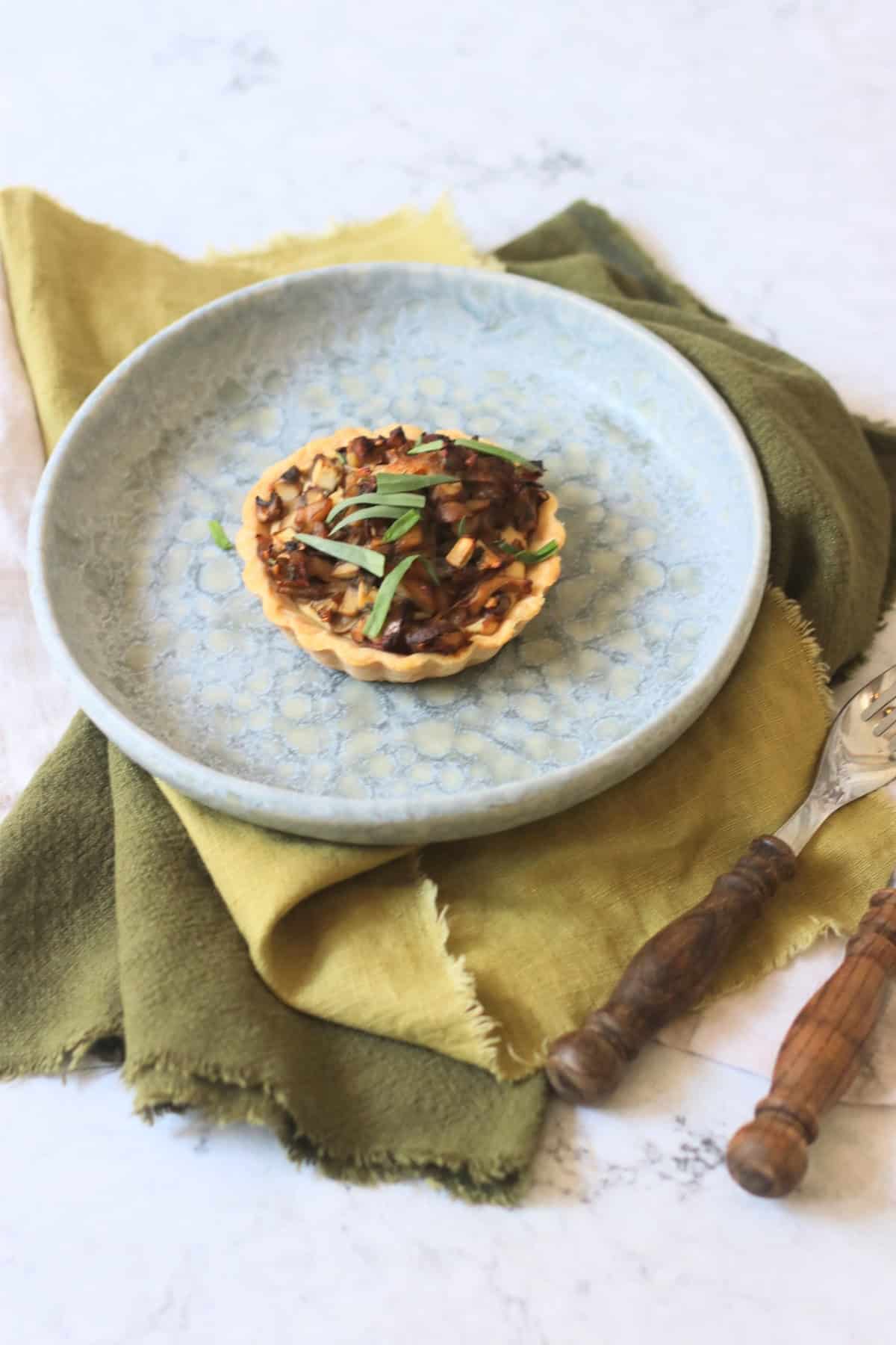 A Vegan Wild Mushroom Tartlet with Caramelised Shallots on a plate with a green napkin and wooden handled knife and fork