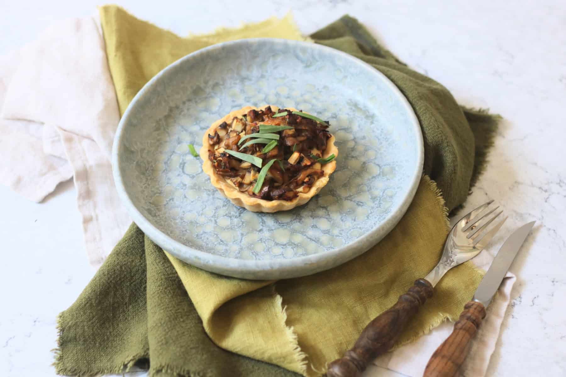 A Vegan Wild Mushroom Tartlet with Caramelised Shallots on a plate with a green napkin and wooden handled knife and fork