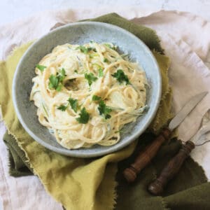 A plate of Vegan Creamy Courgette Linguine topped with fresh parsley served on a blue plate with green napkins and wooden handled knife and fork.