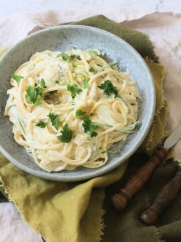 A plate of Vegan Creamy Courgette Linguine topped with fresh parsley served on a blue plate with green napkins and wooden handled knife and fork.