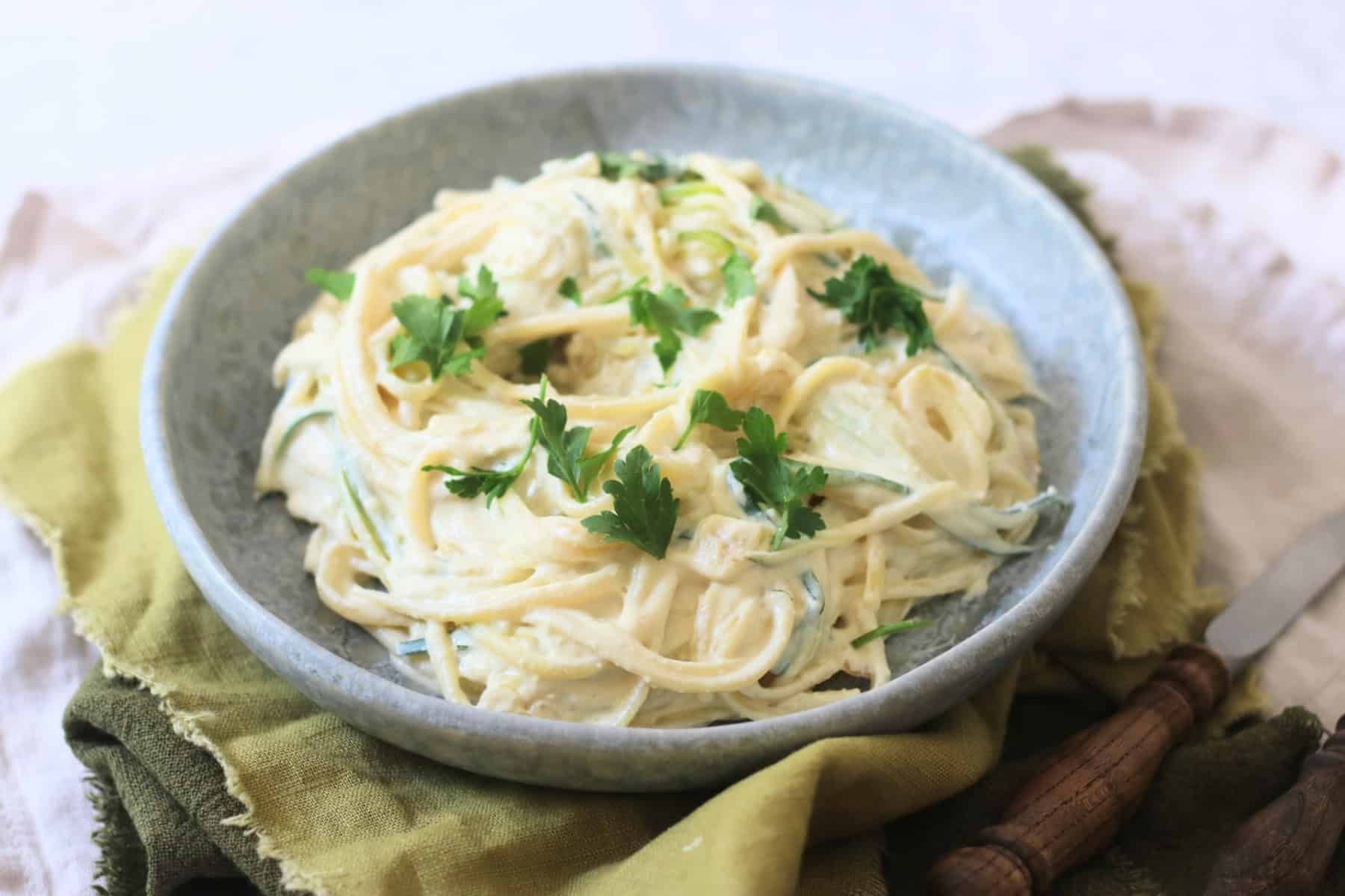 A plate of Vegan Creamy Courgette Linguine topped with fresh parsley served on a blue plate with green napkins and wooden handled knife and fork.