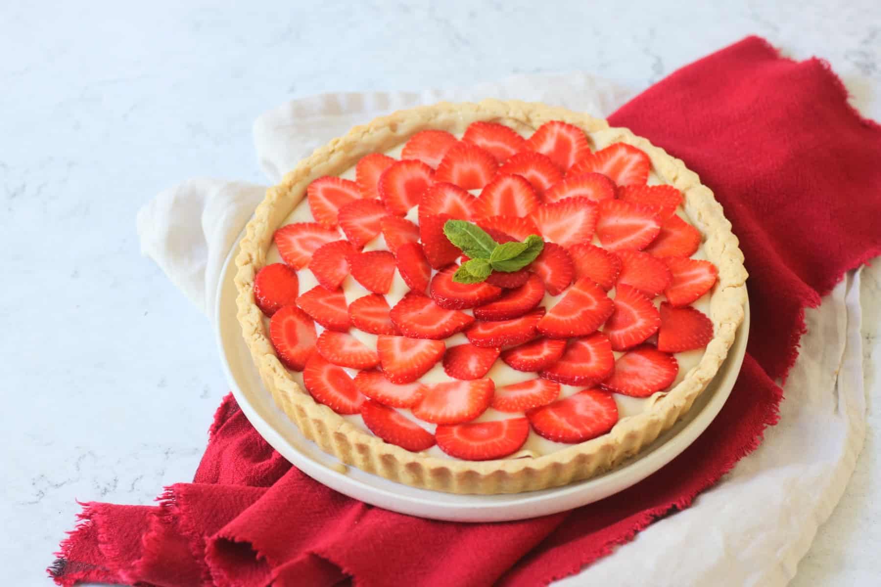 A whole Vegan Strawberry Tart (Vegan Tarte Aux Fraises) made up of a crisp pastry case, vegan creme patisserie and slices of strawberry topped with a couple of mint leaves.