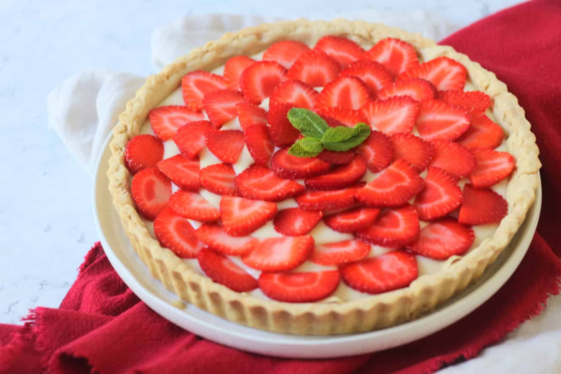 A whole Vegan Strawberry Tart (Vegan Tarte Aux Fraises) made up of a crisp pastry case, vegan creme patisserie and slices of strawberry topped with a couple of mint leaves.