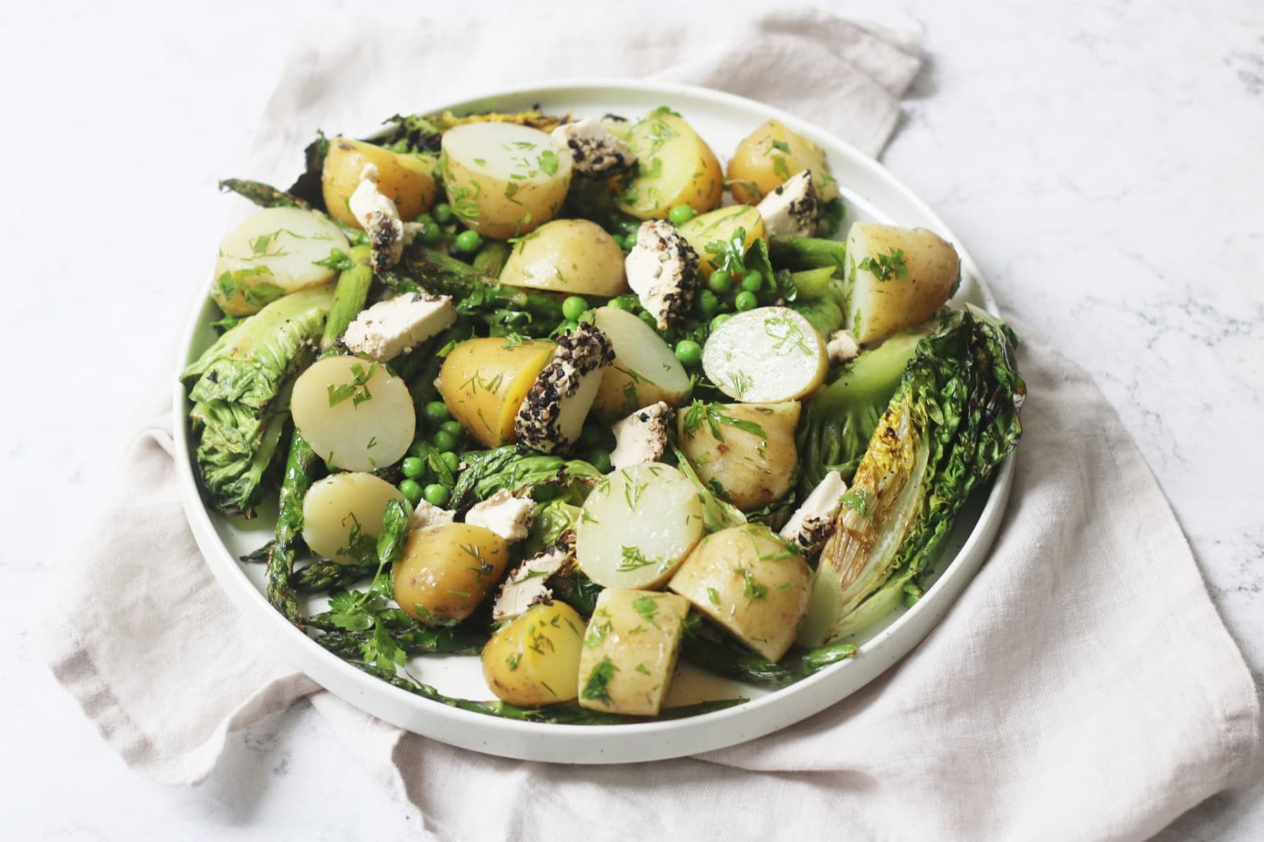 A large plate of BBQ Charred Lettuce Salad with Pepper-Crusted Vegan Goat's Cheese, New Potatoes & Asparagus on a white napkin