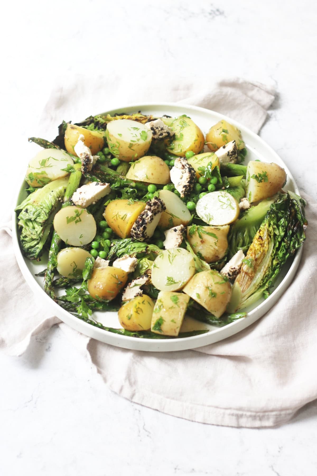 A large plate of BBQ Charred Lettuce Salad with Pepper-Crusted Vegan Goat's Cheese, New Potatoes & Asparagus on a white napkin