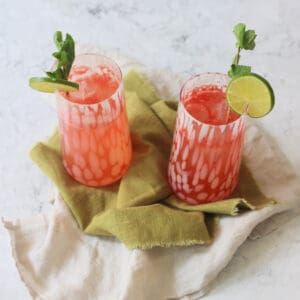 Two tall glasses of Watermelon Agua Fresca garnished with lime wheels and mint served on a green napkin