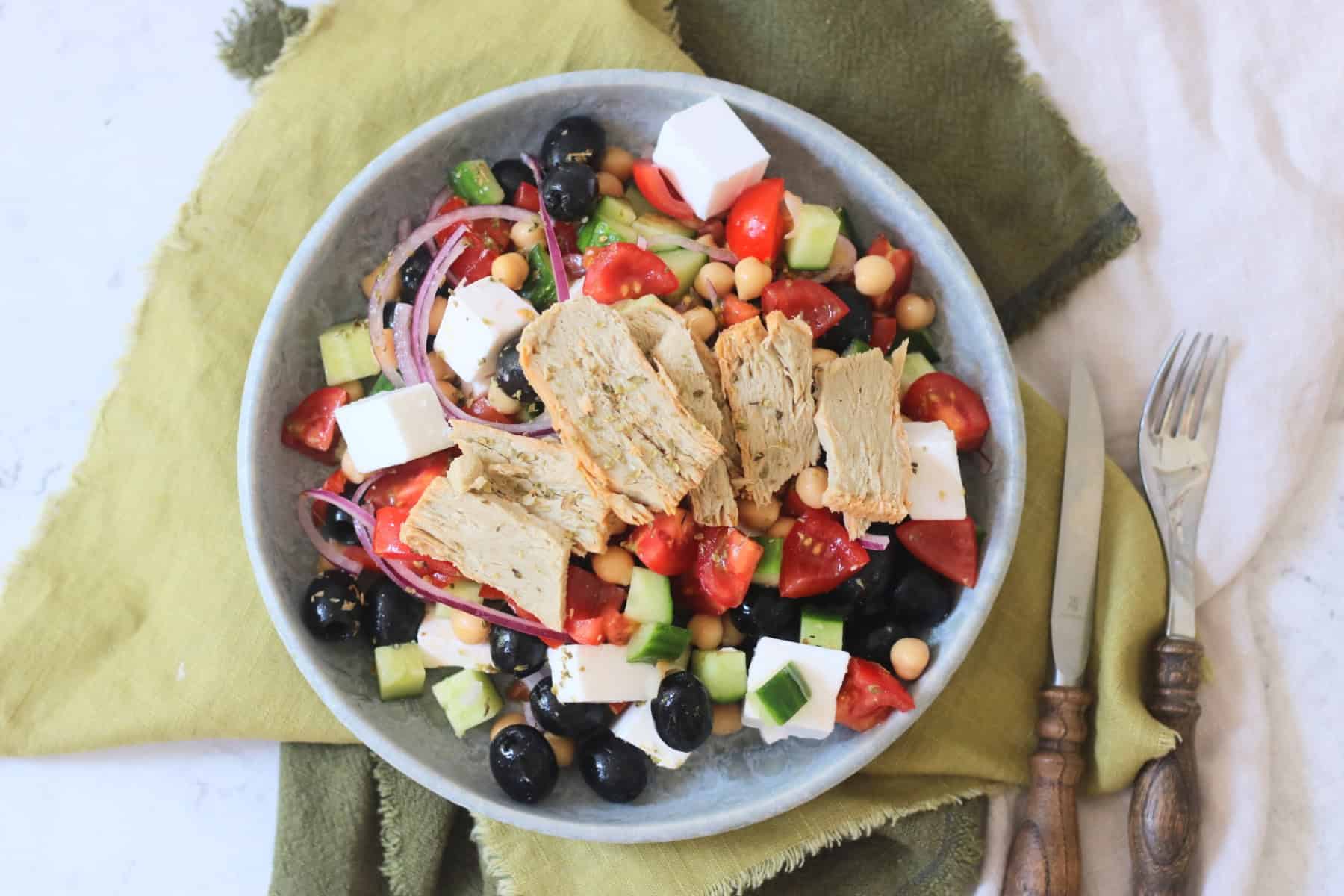 A plate of Greek Inspired Salad with Vegan Chicken Slices sat on green napkins with wooden handled knife and fork.