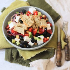 A plate of Greek Inspired Salad with Vegan Chicken Slices sat on green napkins with wooden handled knife and fork.