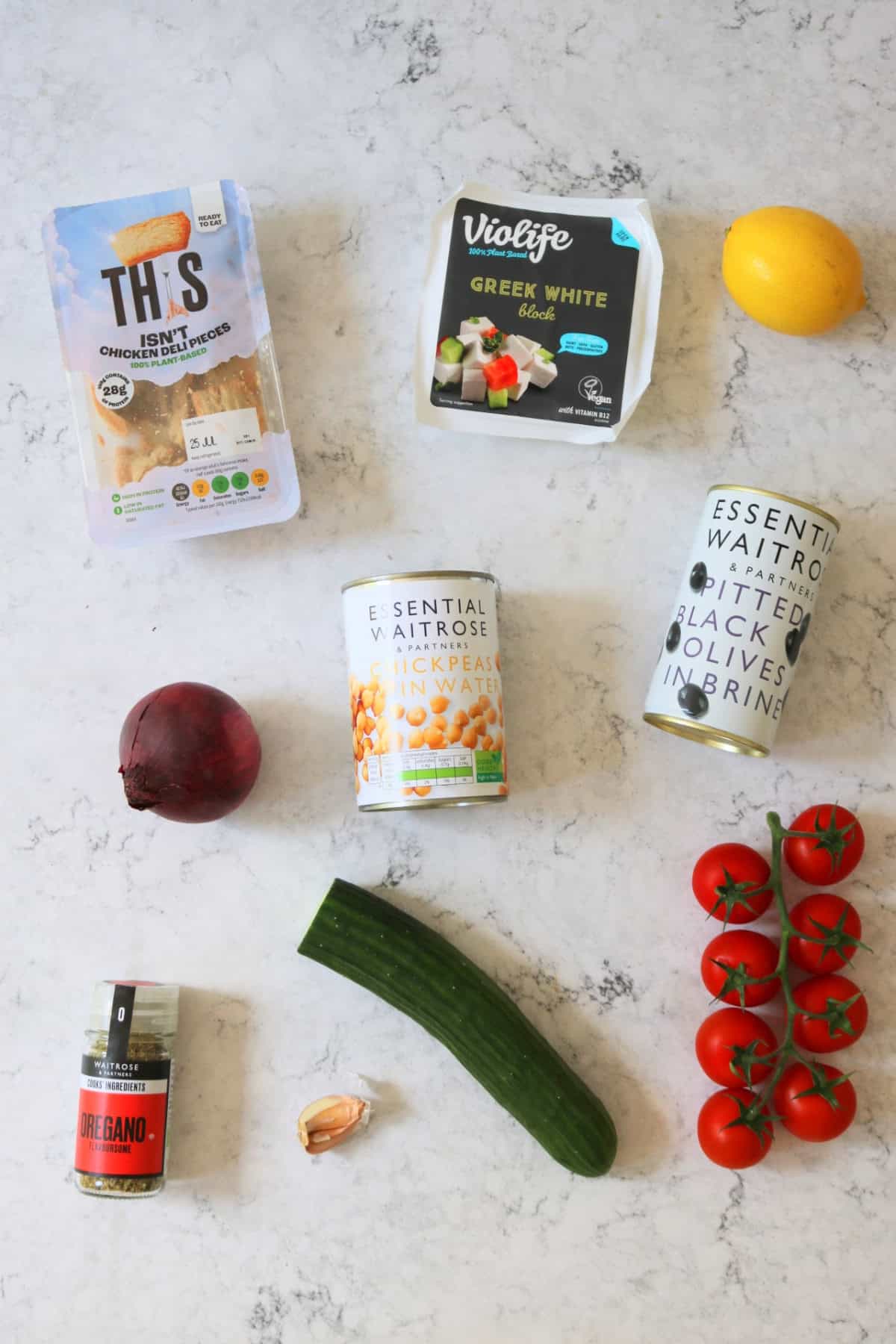 A packet of vegan chicken pieces, a pack of vegan feta, a lemon, a red onion, a tin of chickpeas, a tin of black olives in brine, dried oregano, 2 cloves of garlic, half a cucumber and 8 tomatoes on the vine, all laid out on a white marble counter top.
