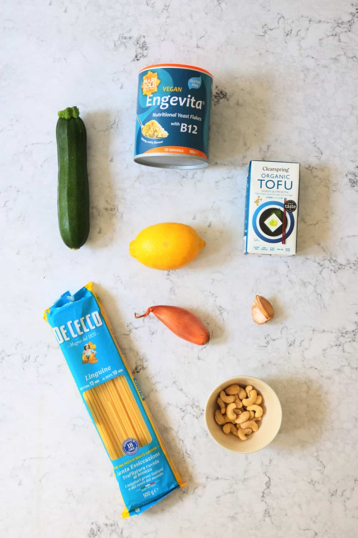 Courgette, nutritional yeast, lemon, silken tofu, linguine, shallot, garlic and cashew nuts laid out on a white marble counter top.