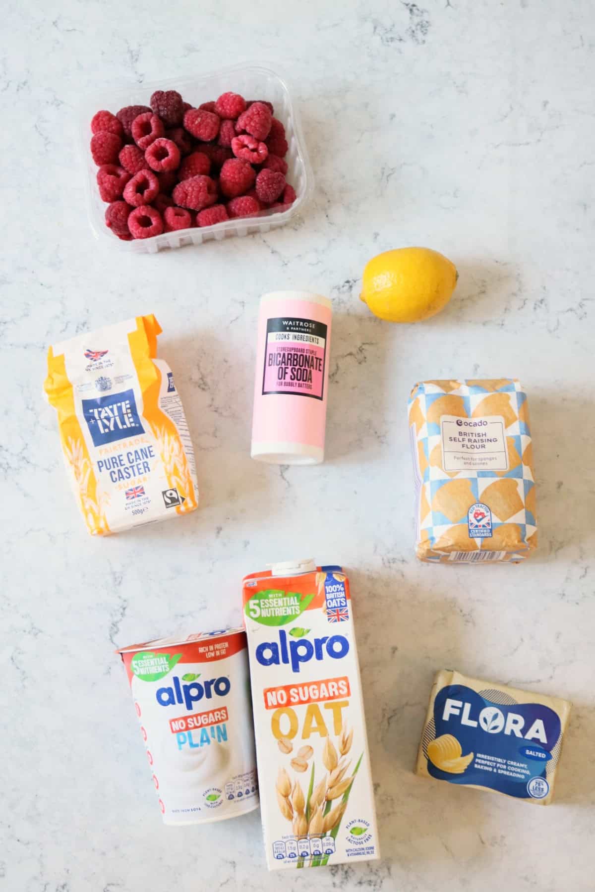 Raspberries, caster sugar, bicarbonate of soda, lemon, self raising flour, dairy-free yogurt, plant milk, vegan butter, all laid out on a white marble counter top
