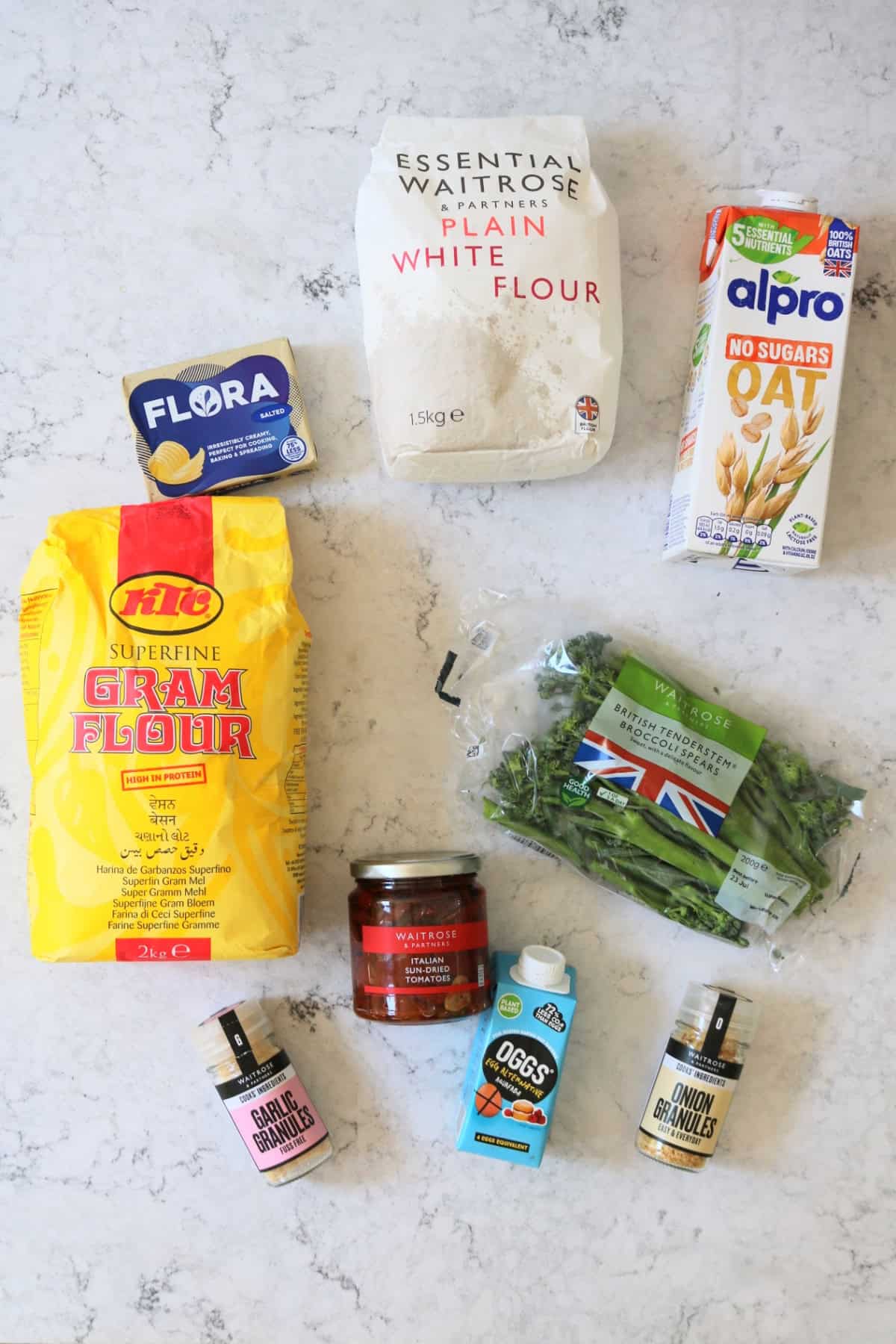 Vegan butter, plain flour, plant milk, chickpea flour, sun-dried tomatoes, Tenderstem broccoli, garlic powder, aquafaba and onion powder laid out on a white marble counter top