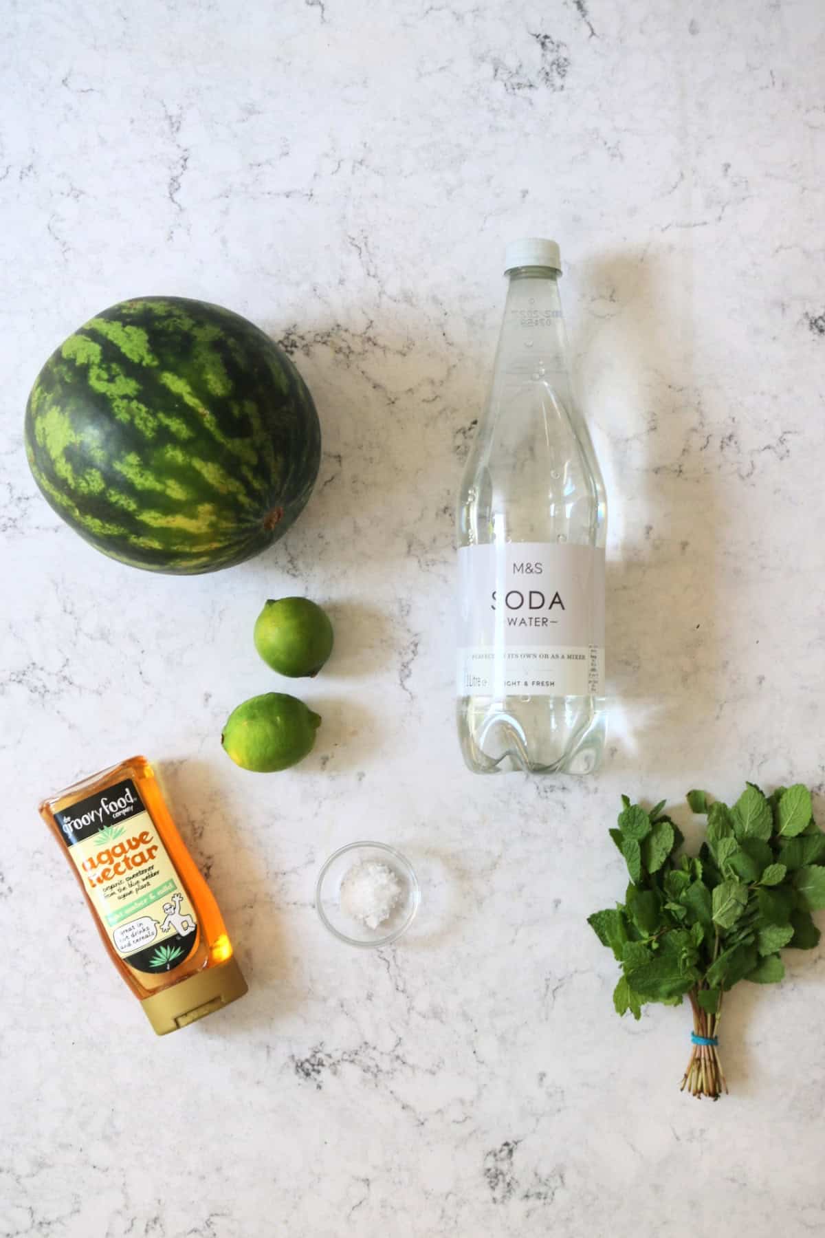 A watermelon, a bottle of soda water, 2 limes, agave nectar, sea salt and mint leaves laid out on a white marble counter top
