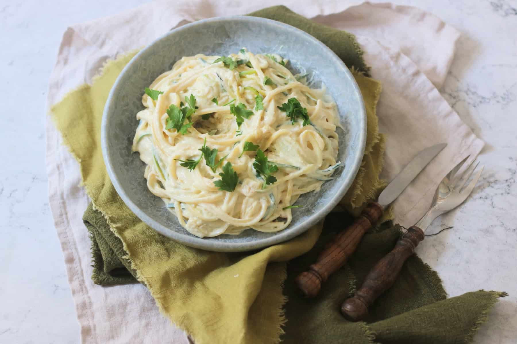 A plate of Vegan Creamy Courgette Linguine served on a blue plate with green napkins and wooden handled knife and fork.