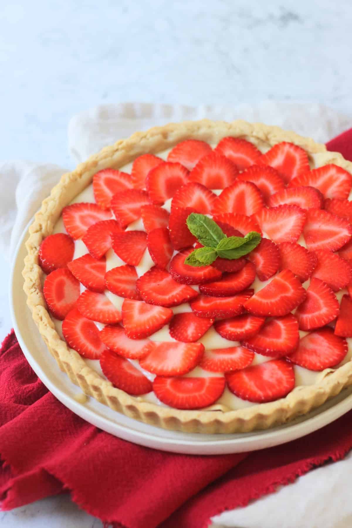 A whole Vegan Strawberry Tart (Vegan Tarte Aux Fraises) made up of a crisp pastry case, vegan creme patisserie and slices of strawberry topped with a couple of mint leaves.