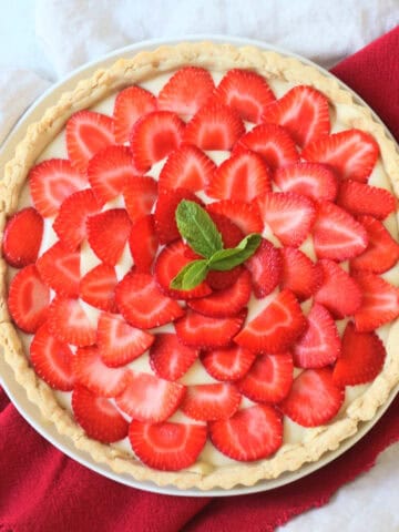 A whole Vegan Strawberry Tart (Vegan Tarte Aux Fraises) made up of a crisp pastry case, vegan creme patisserie and slices of strawberry topped with a couple of mint leaves.