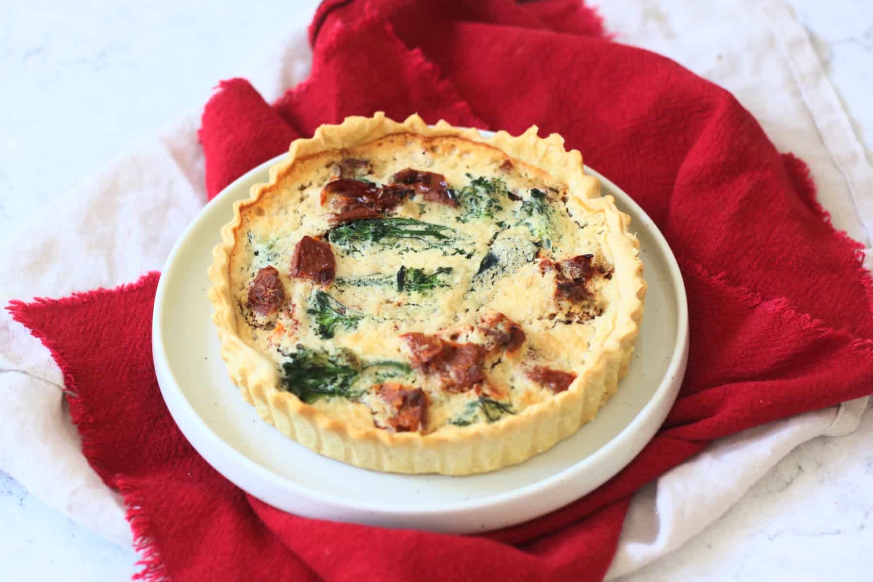A whole Vegan Tenderstem Broccoli and Sun-dried Tomato Quiche on a plate with red and cream napkins