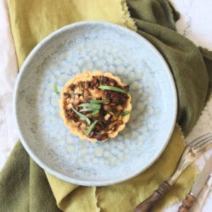 A Vegan Wild Mushroom Tartlet with Caramelised Shallots on a plate with a green napkin and wooden handled knife and fork
