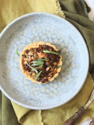 A Vegan Wild Mushroom Tartlet with Caramelised Shallots on a plate with a green napkin and wooden handled knife and fork