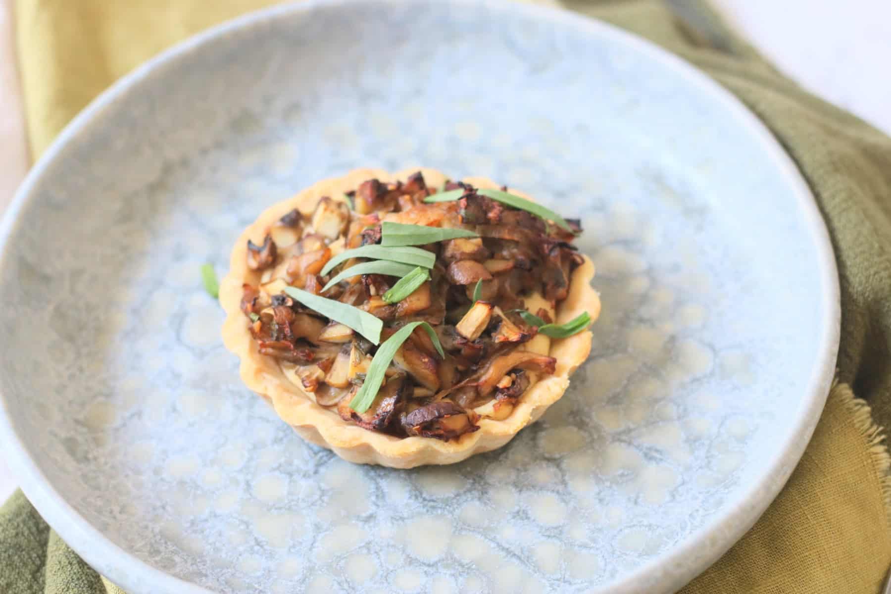 A Vegan Wild Mushroom Tartlet with Caramelised Shallots on a plate with a green napkin and wooden handled knife and fork