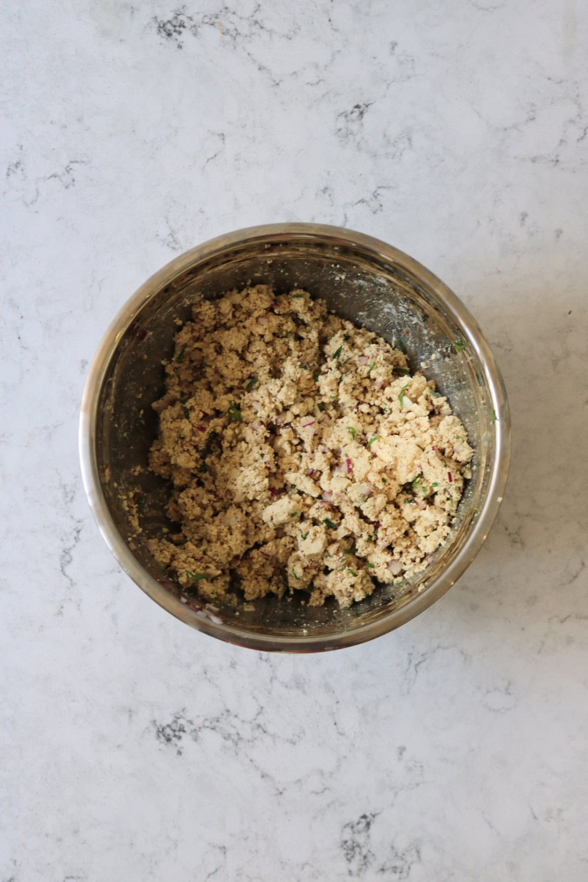 Crumbled tofu mixed with herbs, spices, diced red onion and chickpea flour in a metal mixing bowl
