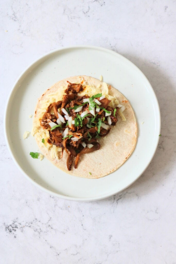 A corn tortilla filled with vegan cheese and shredded oyster mushrooms and jackfruit, topped with diced white onion and coriander