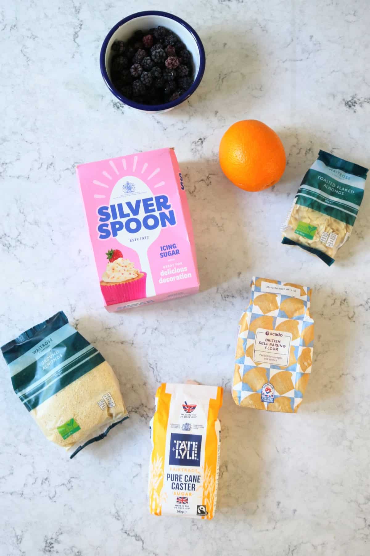 Blackberries, icing sugar, orange, ground almonds, self raising flour, caster sugar all laid out on a white marble counter top