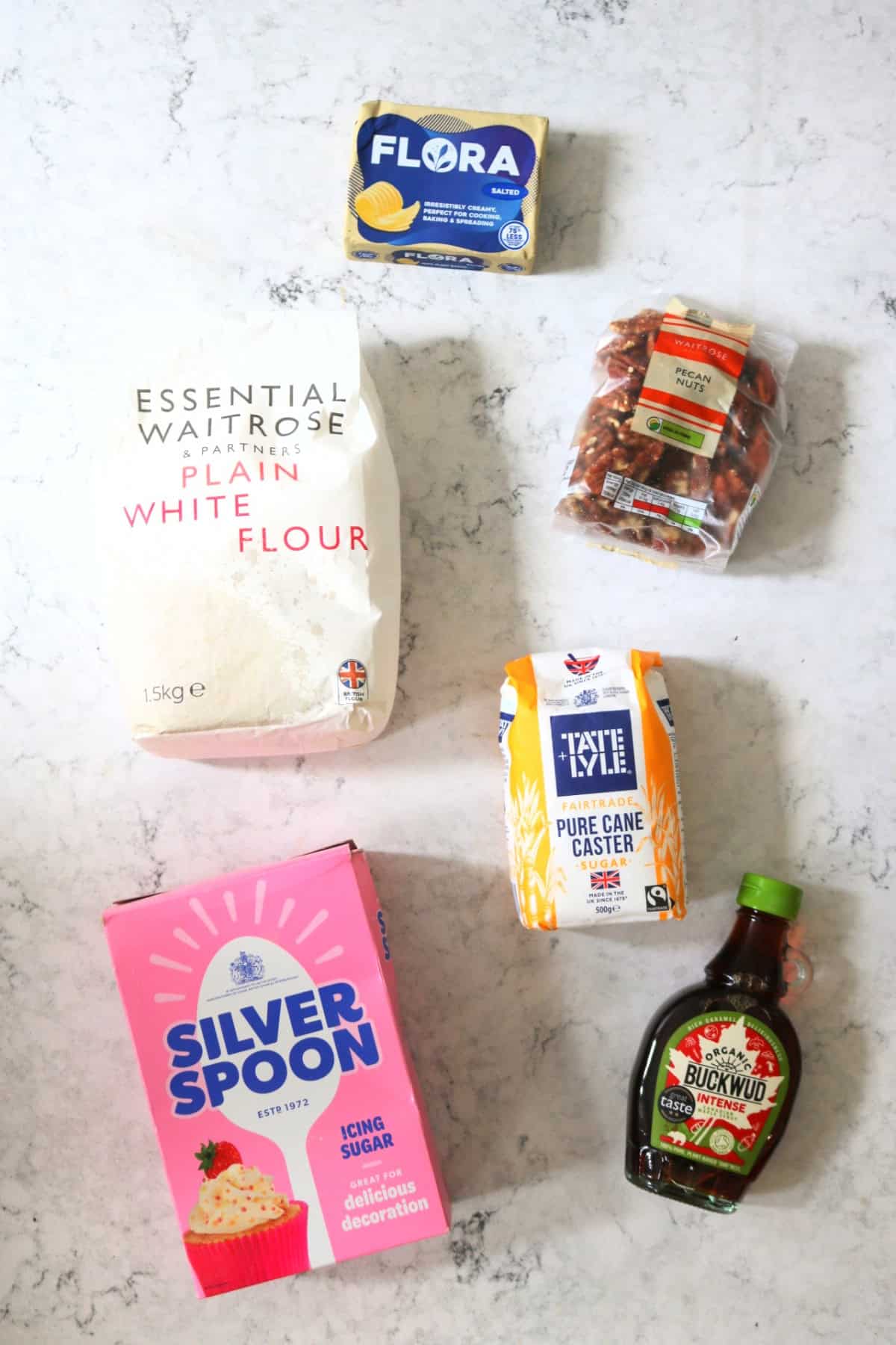 Vegan butter, plain flour, pecans, caster sugar, icing sugar, maple syrup all laid out on a white marble counter top.