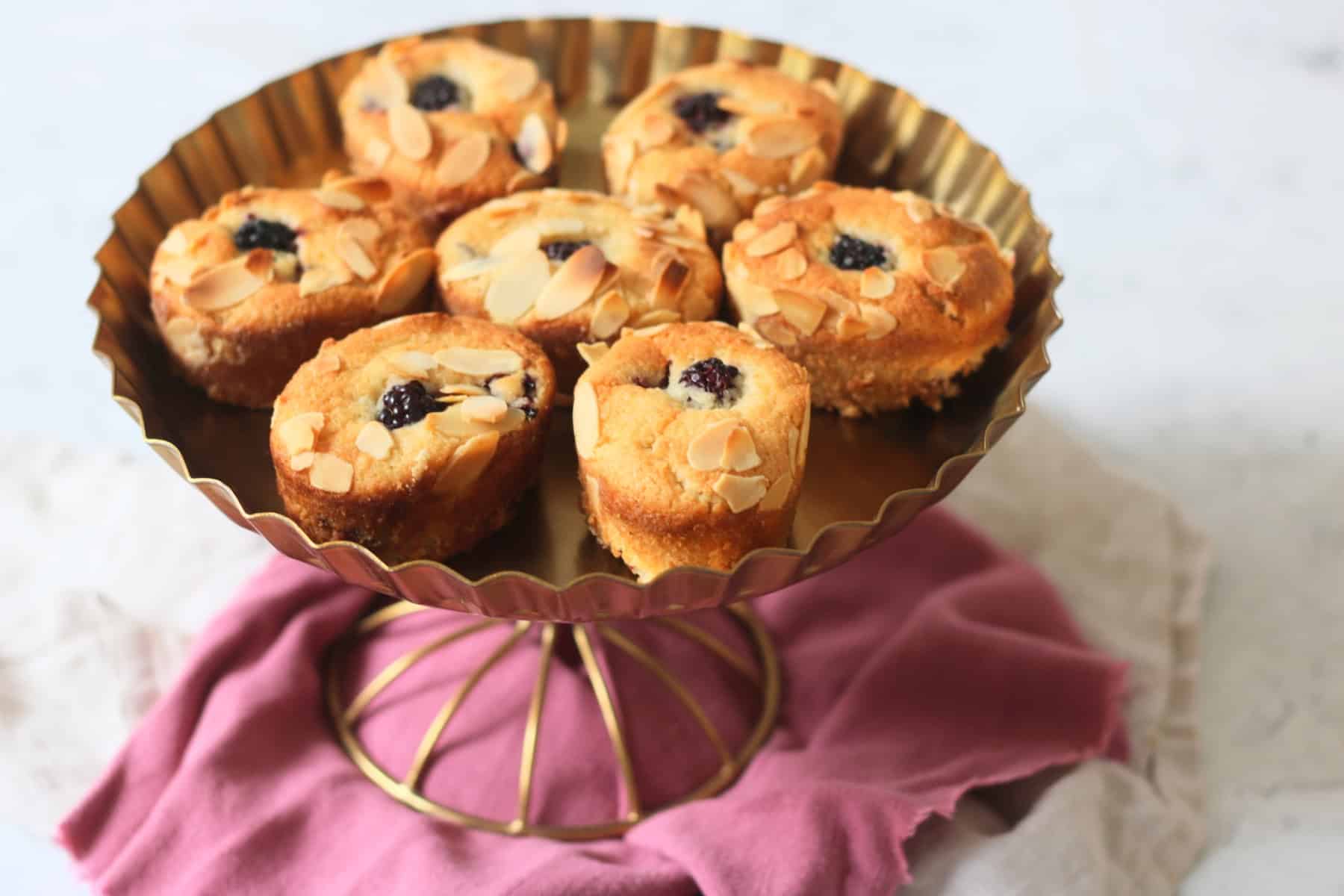 Seven Vegan Blackberry and Orange Friands on a gold cake stand sat on top of a pink and white napkin