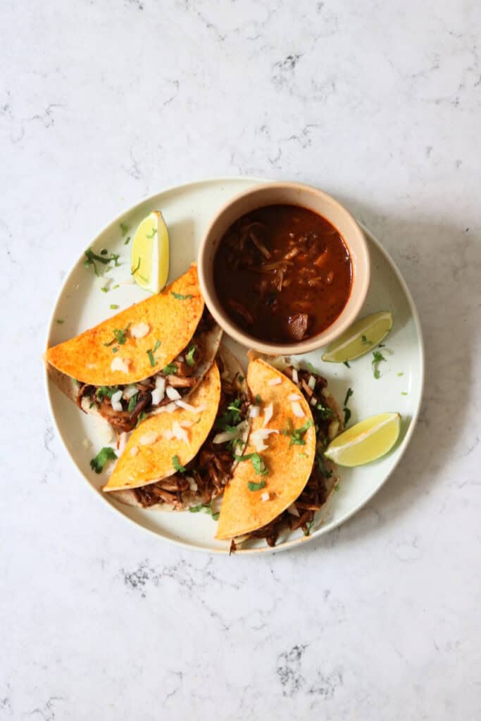 Oyster mushroom and jackfruit stuffed Vegan Birria Tacos topped with diced white onion and coriander and served with wedges of lime and cooking liquor to dip