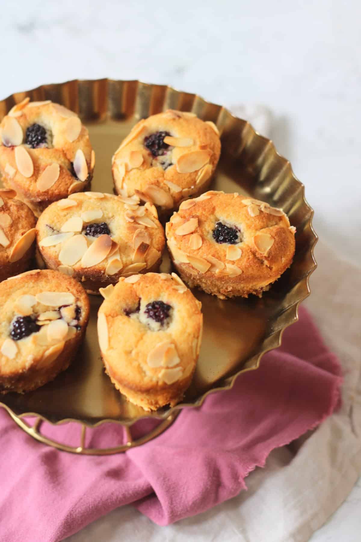 Seven Vegan Blackberry and Orange Friands on a gold cake stand sat on top of a pink and white napkin