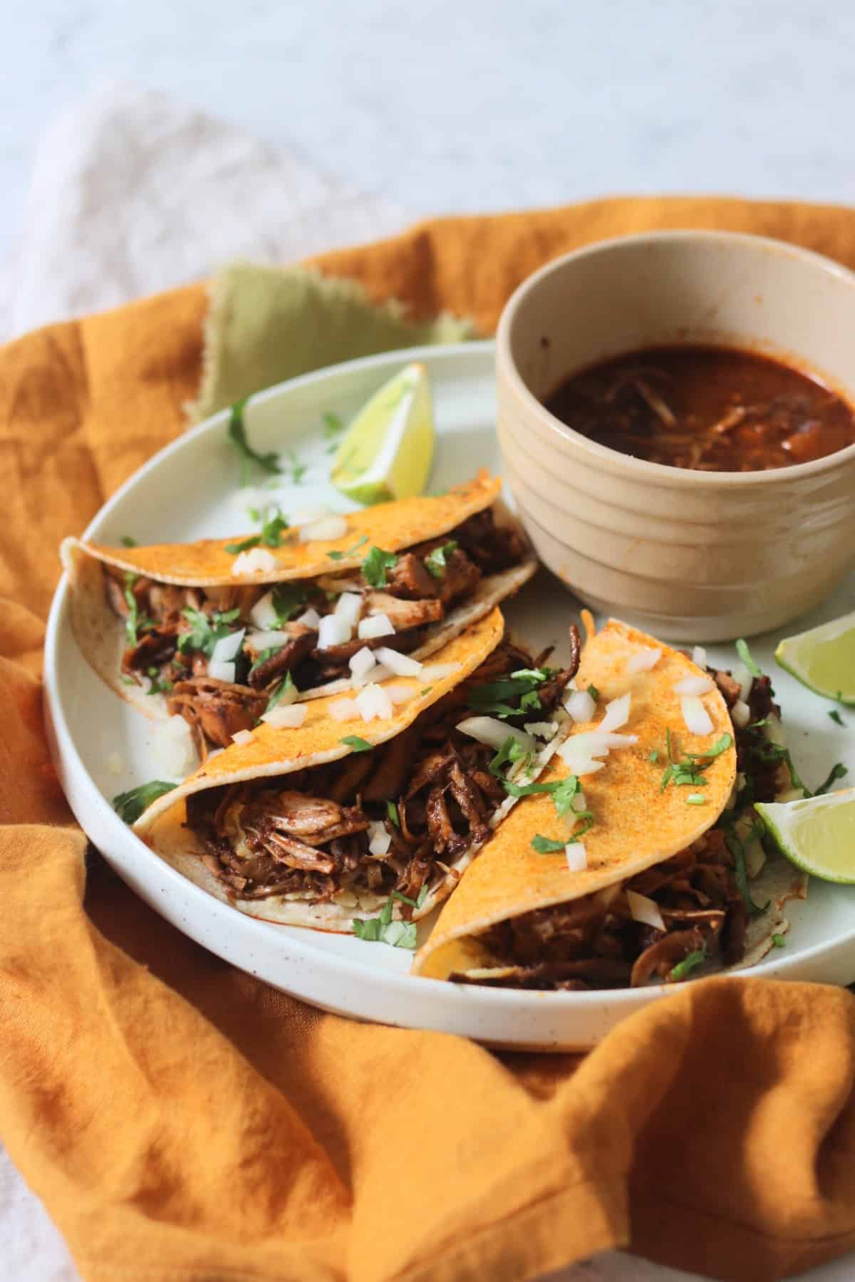 Oyster mushroom and jackfruit stuffed Vegan Birria Tacos topped with diced white onion and coriander and served with wedges of lime and cooking liquor to dip