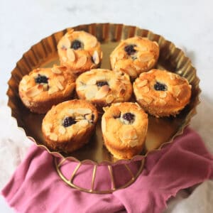Seven Vegan Blackberry and Orange Friands on a gold cake stand sat on top of a pink and white napkin