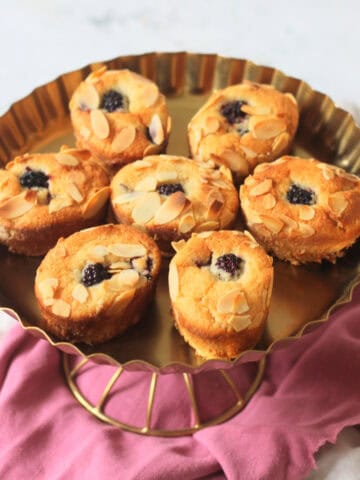 Seven Vegan Blackberry and Orange Friands on a gold cake stand sat on top of a pink and white napkin
