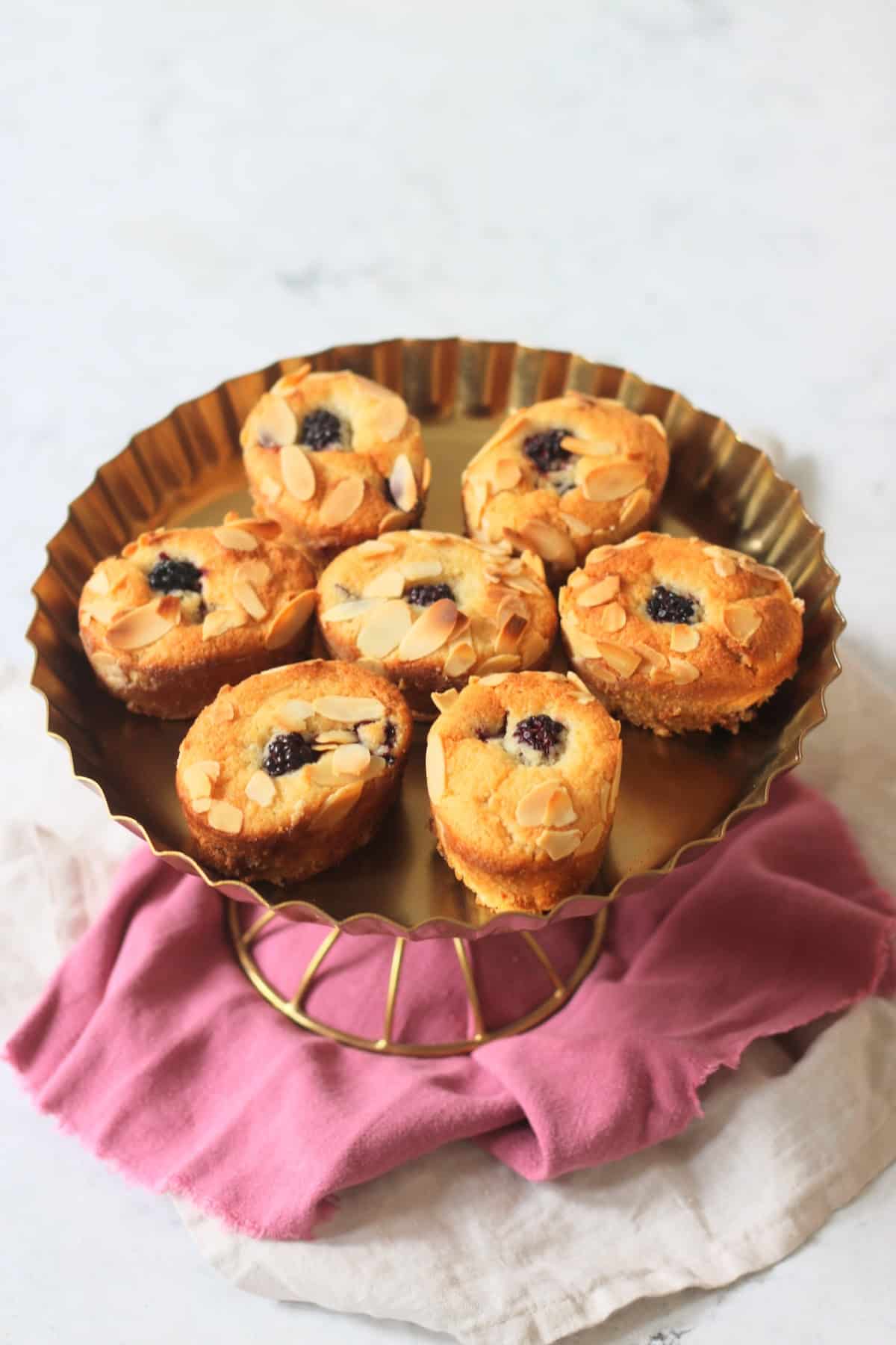Seven Vegan Blackberry and Orange Friands on a gold cake stand sat on top of a pink and white napkin