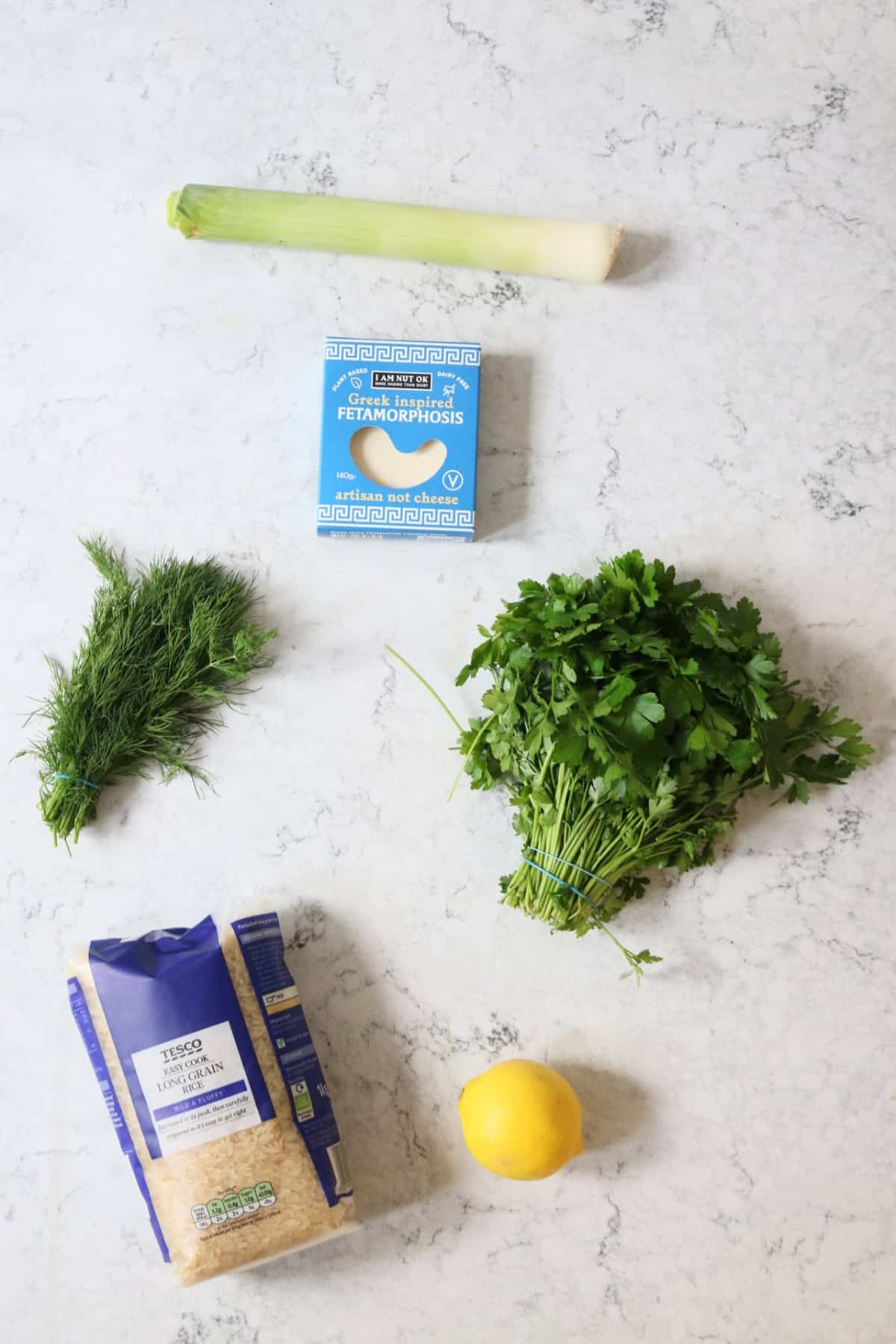 A leek, vegan feta, fresh dill, fresh parsley, rice and lemon laid out on a white marble countertop