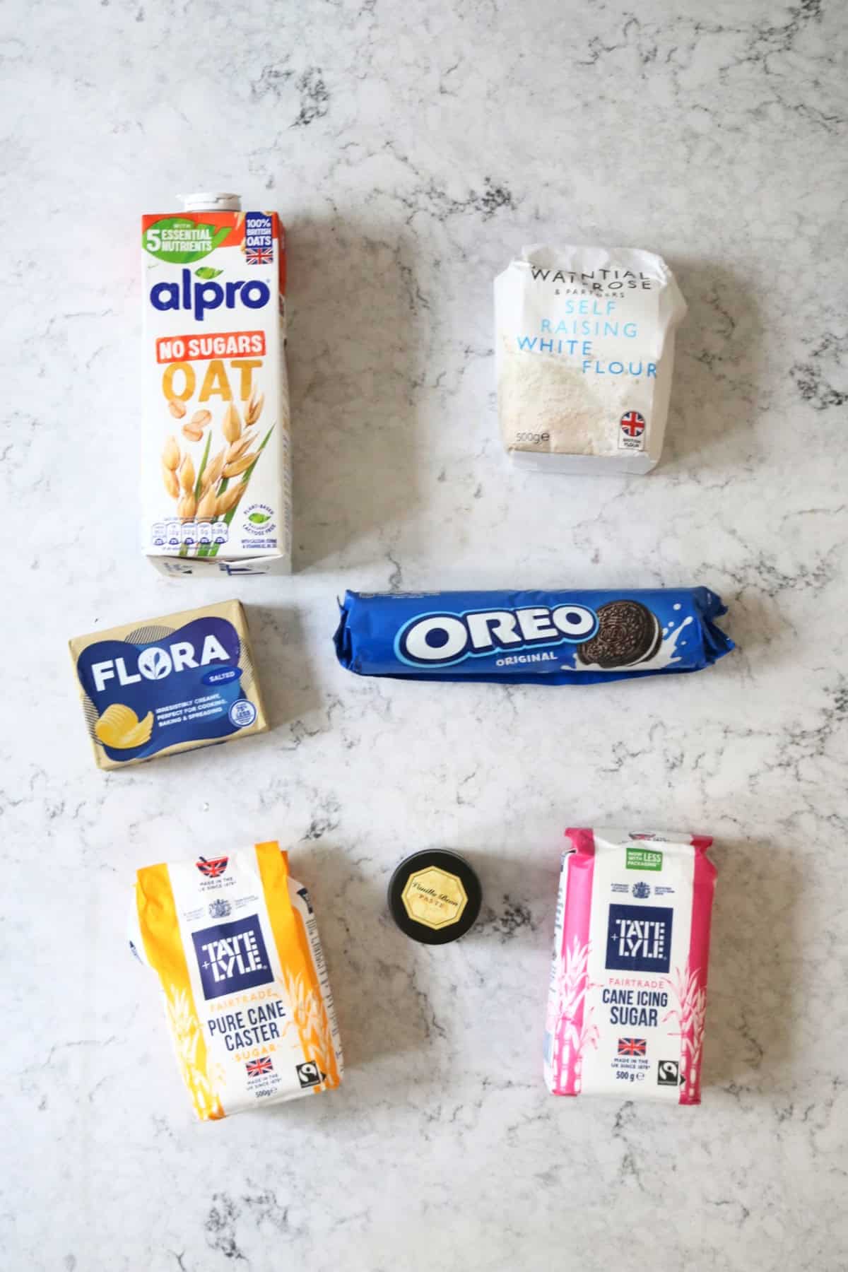 Plant milk, self raising flour, vegan butter, Oreos, caster sugar, vanilla paste and icing sugar laid out on a white marble counter top