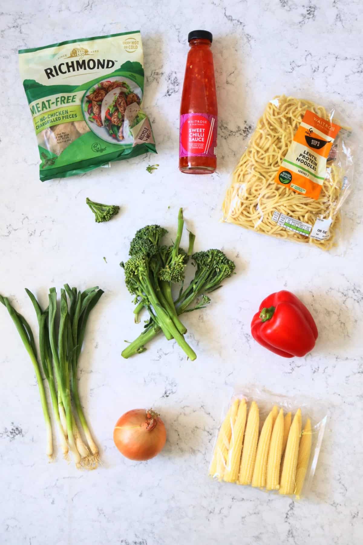 Vegan chicken pieces, sweet chilli sauce, fresh vegan noodles, tenderstem broccoli, red pepper, spring onions, white onions and baby sweetcorn laid out on a white counter top