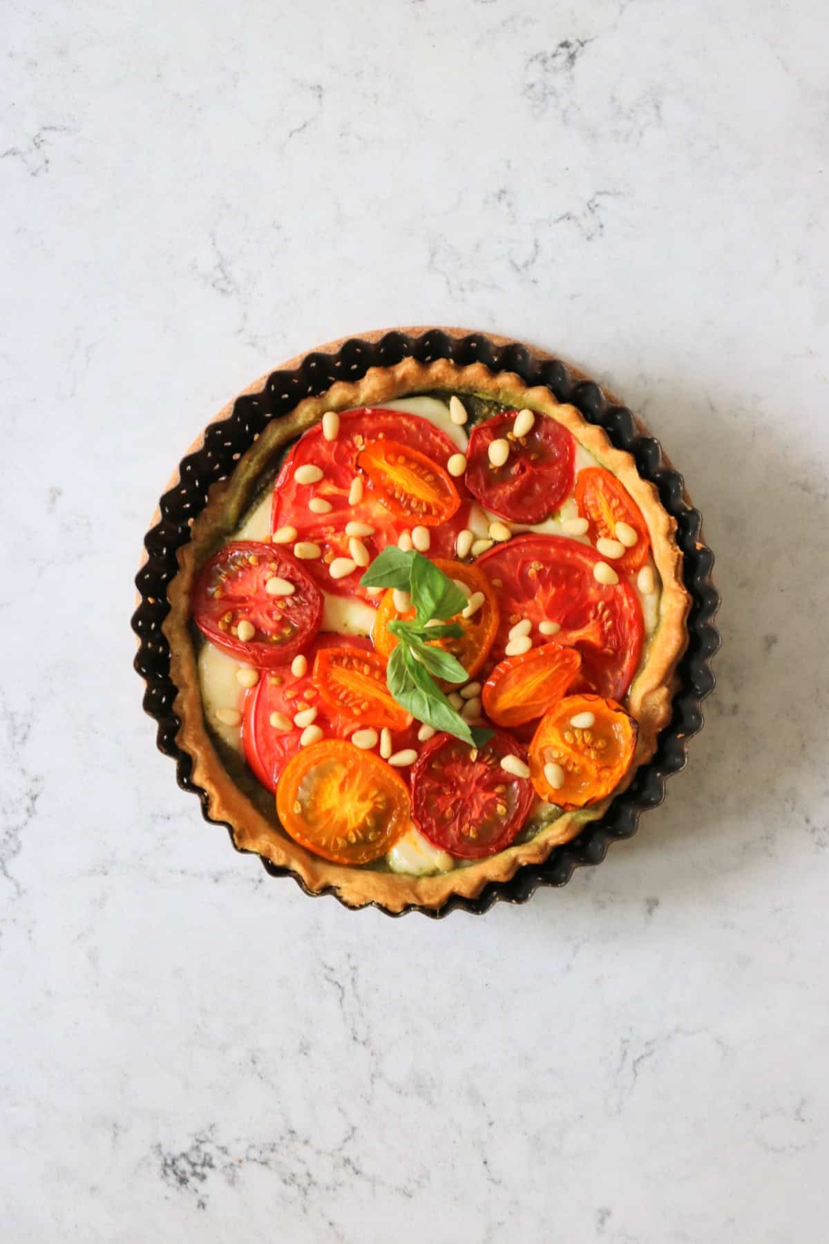 Vegan Tomato Mozzarella and Pesto Picnic Tart topped with pine nuts and basil, fresh out of the oven and still in the tin