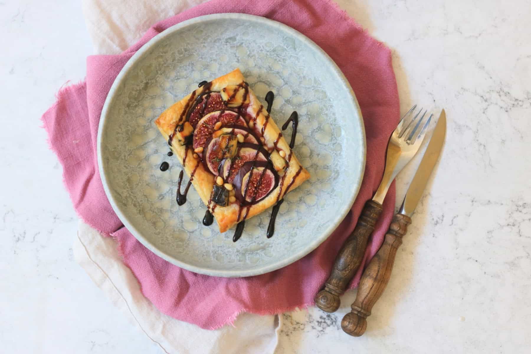 A Vegan Fig and Blue Cheese Tart on a blue plate, drizzled with balsamic glaze and served with a pink napkin and wooden handled cutlery